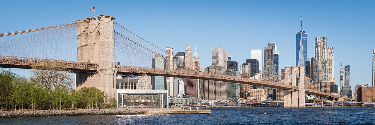 The Brooklyn Bridge and the Lower Manhattan skyline seen from Pebble Beach; the glass pavilion of Jane's Carousel sits below the bridge, with One World Trade Ce
